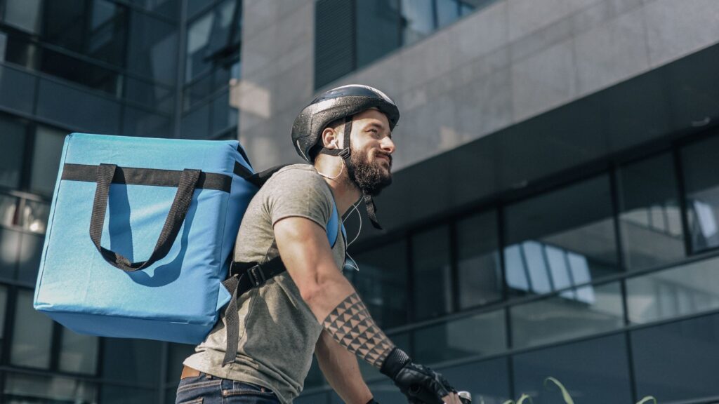 A smiling food delivery man on a bike carrying an insulated box on his back.
