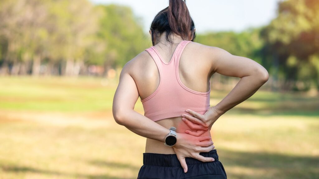 Woman in activewear at a park holding her lower back in pain. 