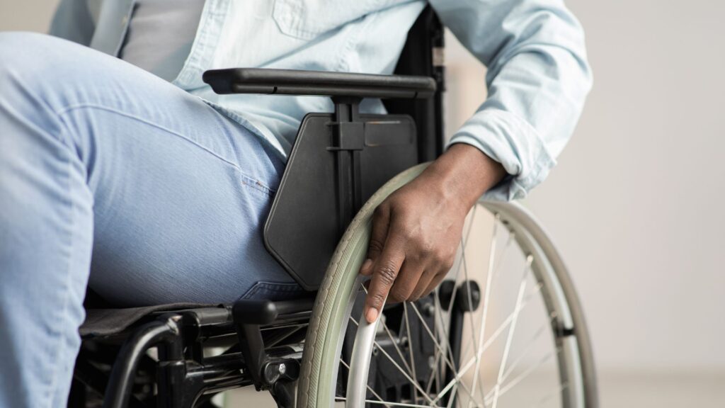 A close-up shot of a man wearing blue jeans and a light blue buttoned shirt in his wheelchair.