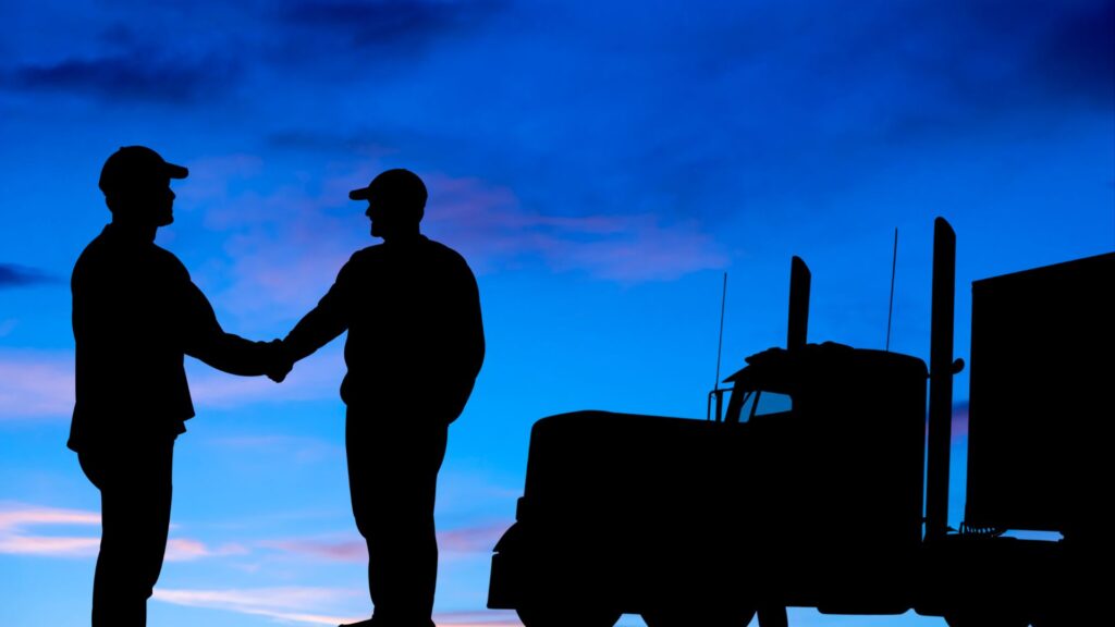 Silhouette of two truck drivers shaking hands next to a big rig truck, sunset sky in the background.